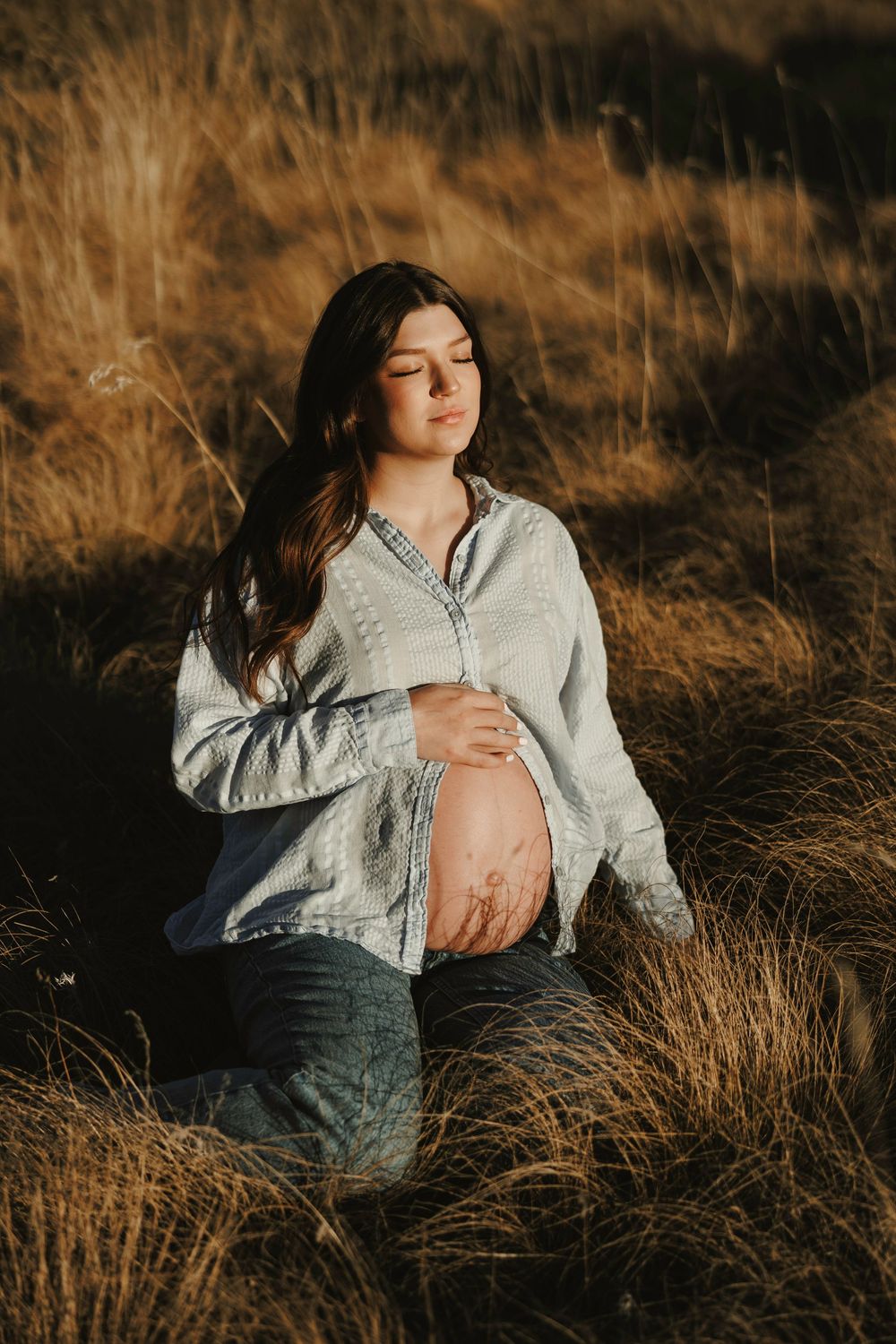 Pregnant woman sits peacefully in golden grass field at sunset, cradling bare belly in open linen shirt.