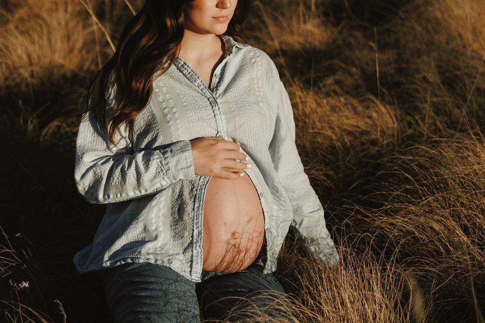 Close-up of pregnant womans bare belly cradled gently in warm golden sunlight among tall dry grasses.
