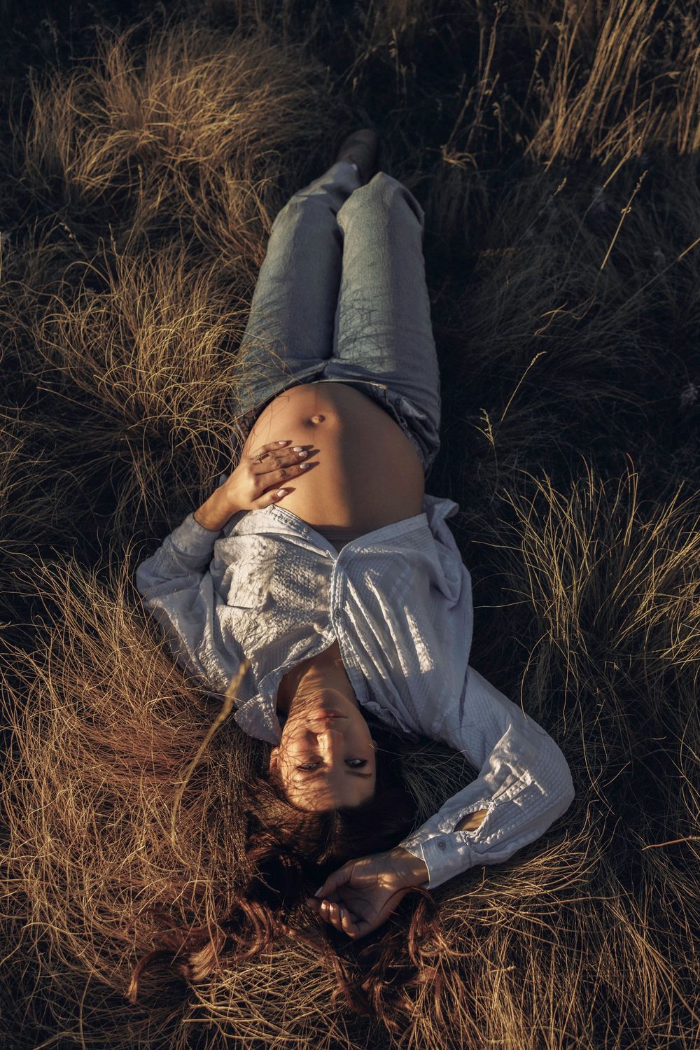 Pregnant woman lies in golden grass at sunset, open shirt revealing belly during outdoor maternity shoot.