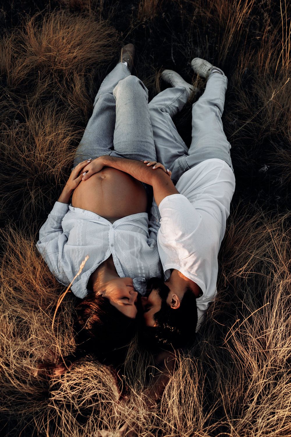Couple lies together in golden grass, man tenderly embracing pregnant partners belly during maternity session.