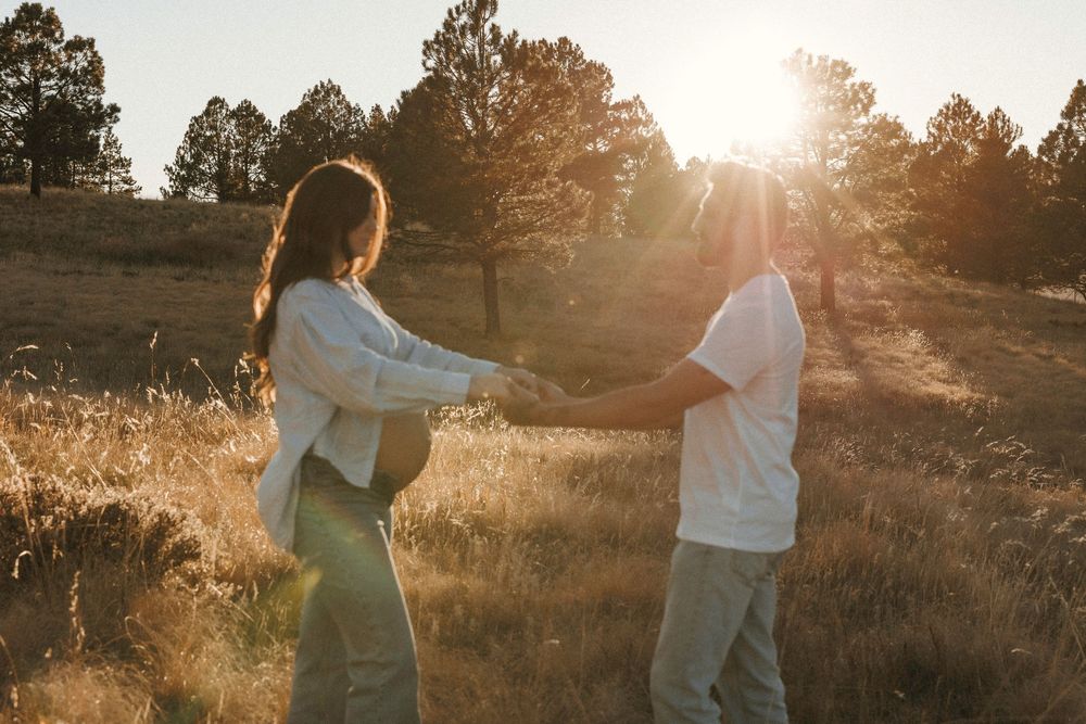 Expecting couple holding hands in sunlit golden meadow during romantic outdoor maternity photoshoot.