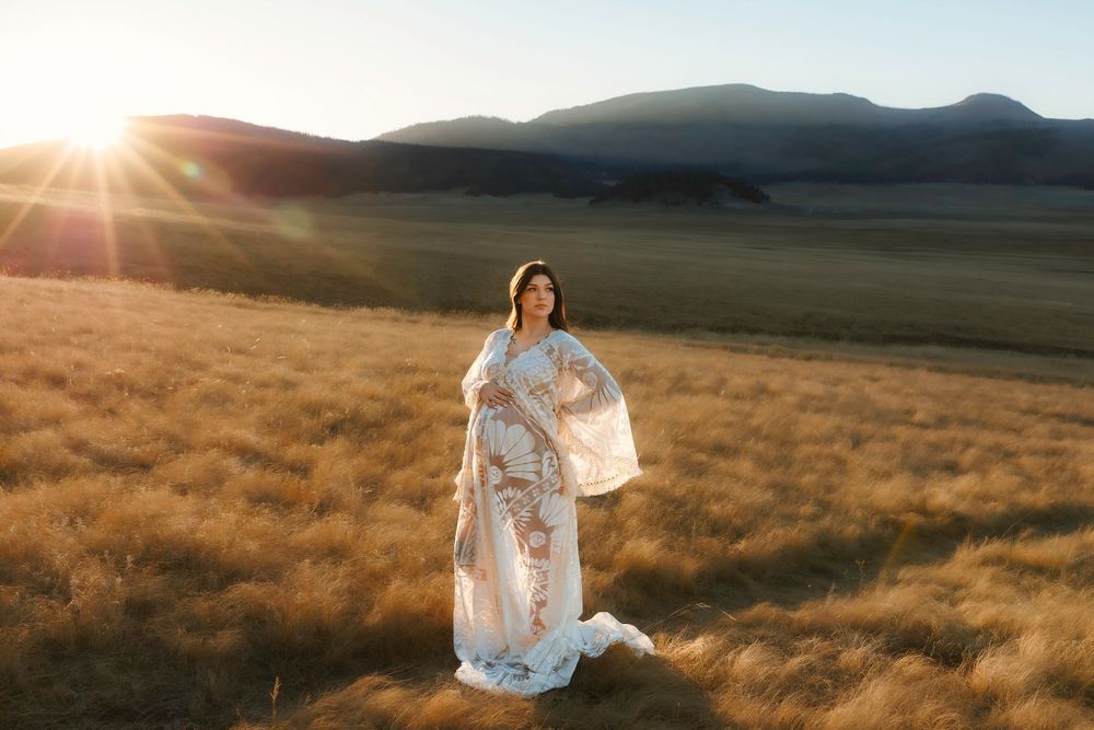 Albuquerque maternity session in flowing white lace client closet dress standing in vast golden field with mountains at sunset.