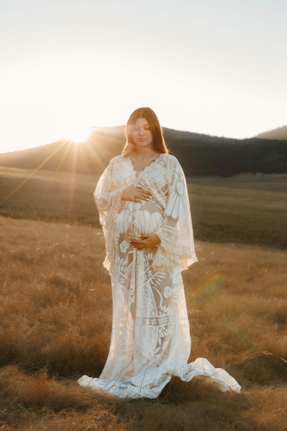 Pregnant woman in white lace boho gown cradling belly in golden field at sunset with mountain backdrop.