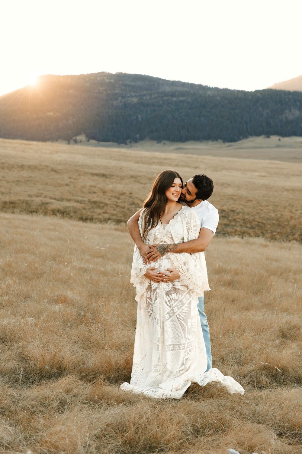Loving couple embracing in golden field at sunset, man kissing pregnant woman in white lace gown.