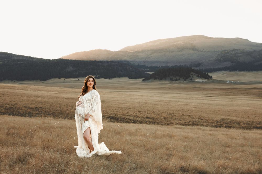 Pregnant woman in white lace dress walking through open golden field with rolling mountains behind.