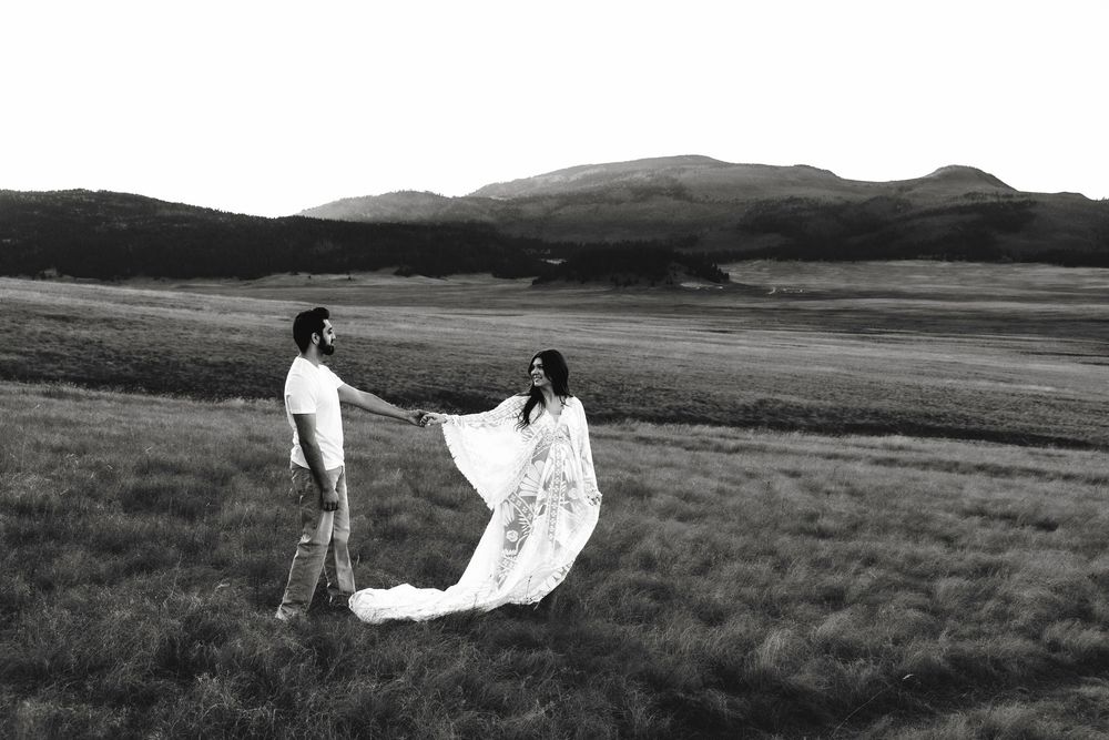 Black and white photo of couple holding hands in open field, pregnant woman in flowing lace gown.