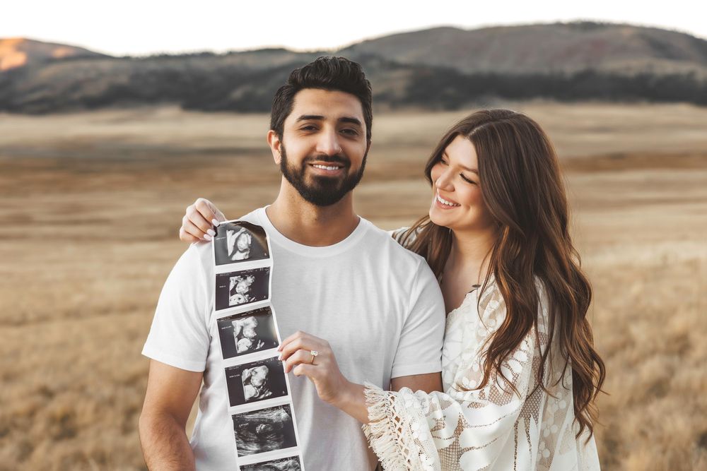 Smiling couple holding ultrasound strip photos during outdoor maternity session in golden meadow.