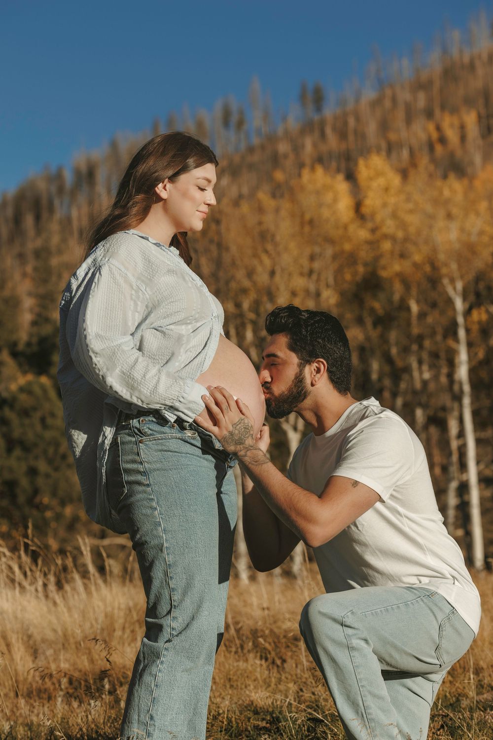 Man kneels to kiss pregnant womans bare belly in autumn field with colorful fall foliage behind them.