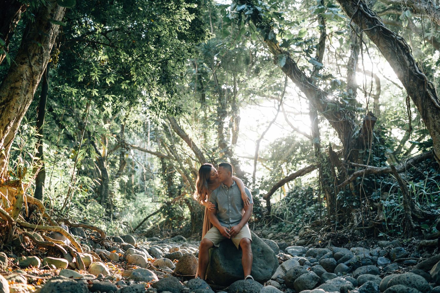 A couple poses together on rocky terrain in a sunlit wooded area.