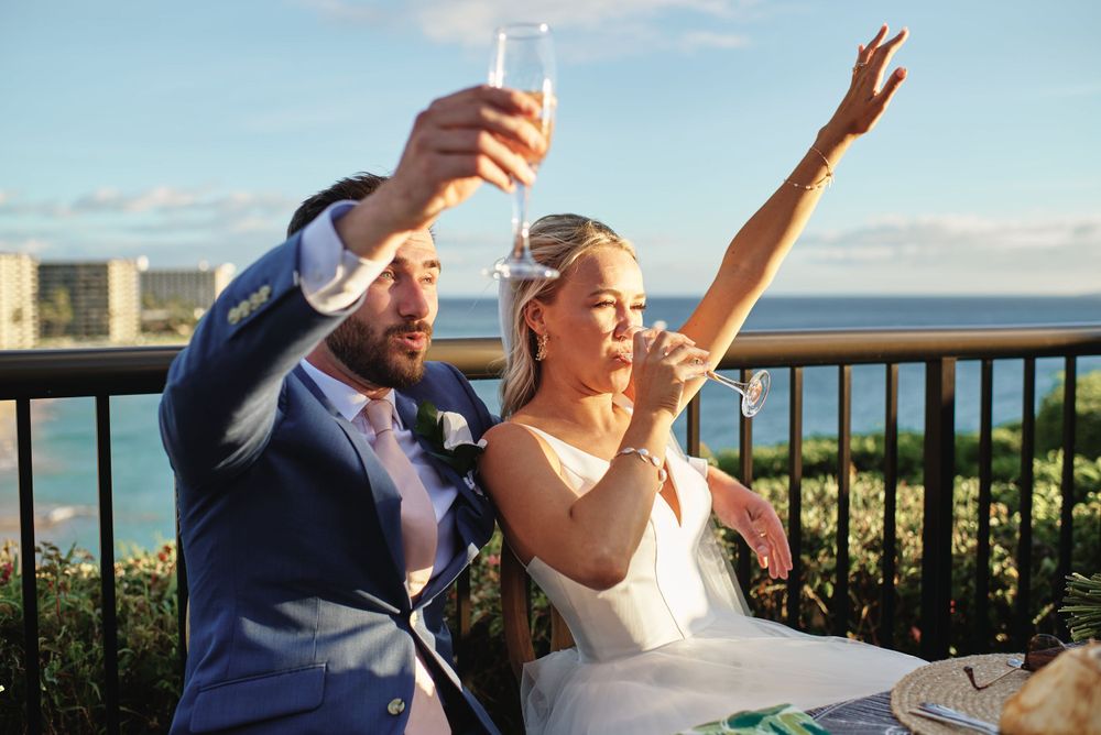 Newlyweds celebrate with raised glasses on oceanfront balcony.