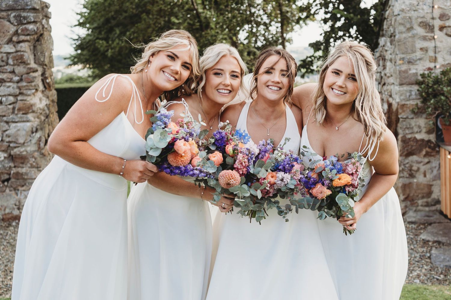 Bride and bridesmaids smiling together outdoors, holding colourful wedding bouquets during a South Wales summer wedding.