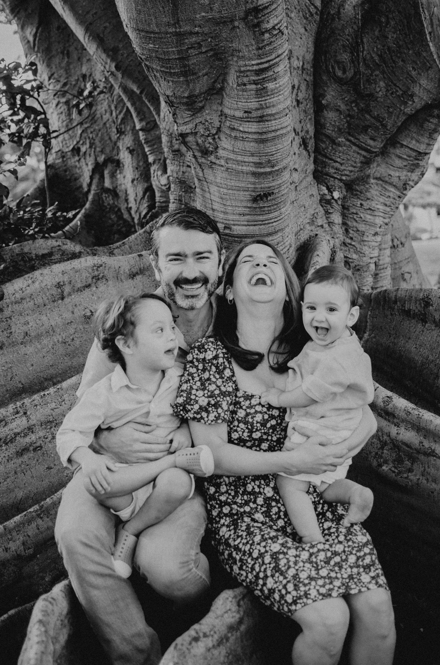 A happy family shares genuine laughter while sitting together on large tree roots in black and white.