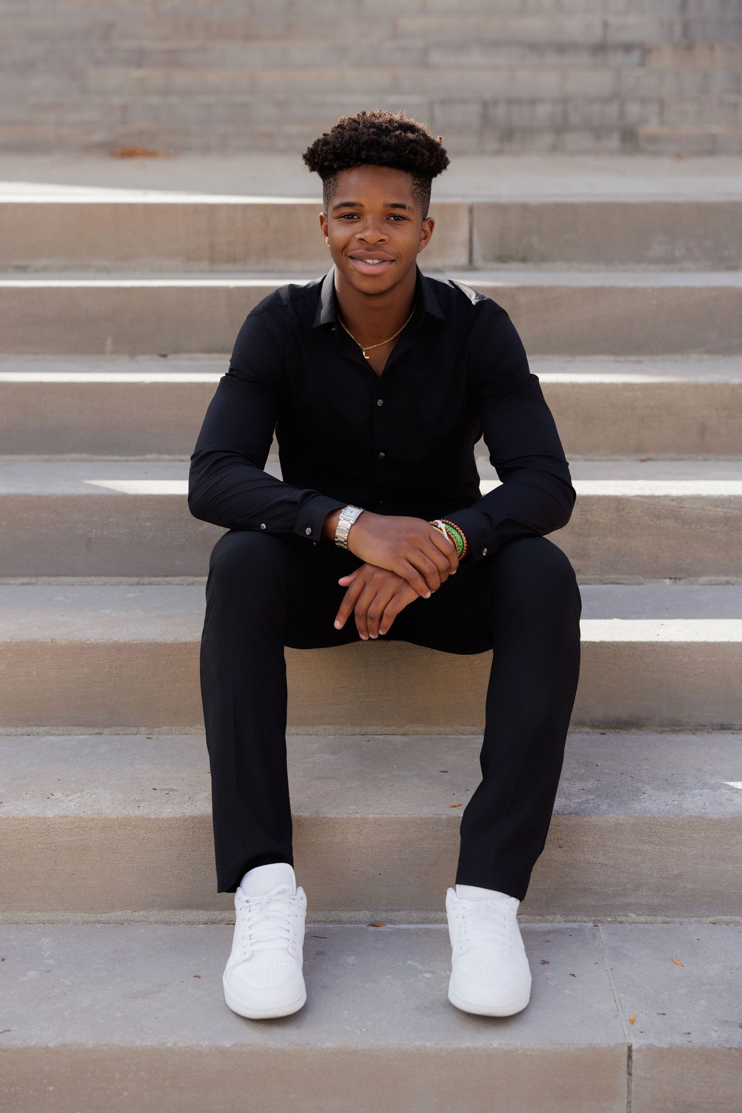 A person in all black attire sitting casually on concrete steps wearing white sneakers.