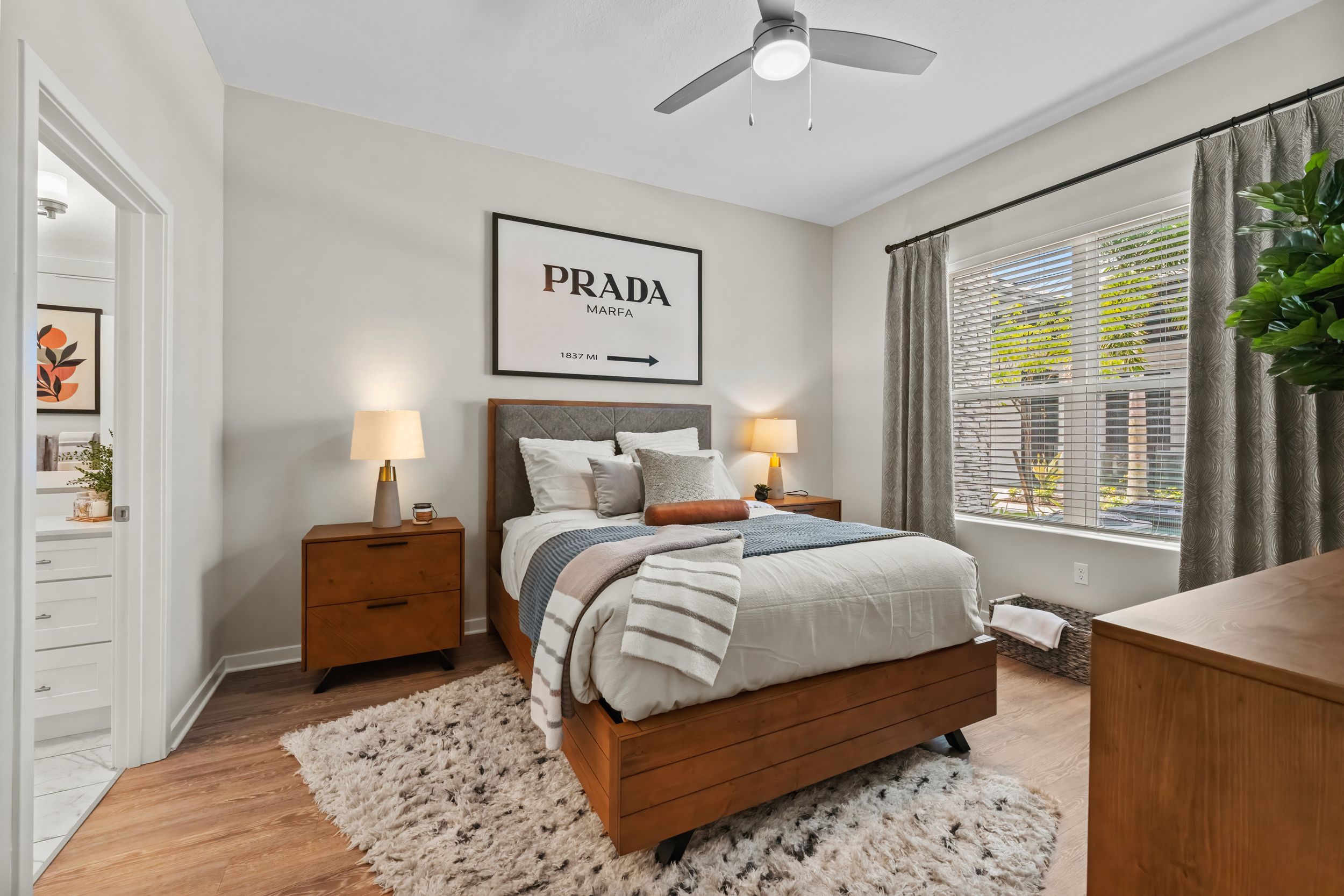 Modern bedroom with Prada artwork, wood furniture, plush rug, ceiling fan and natural light from large window.