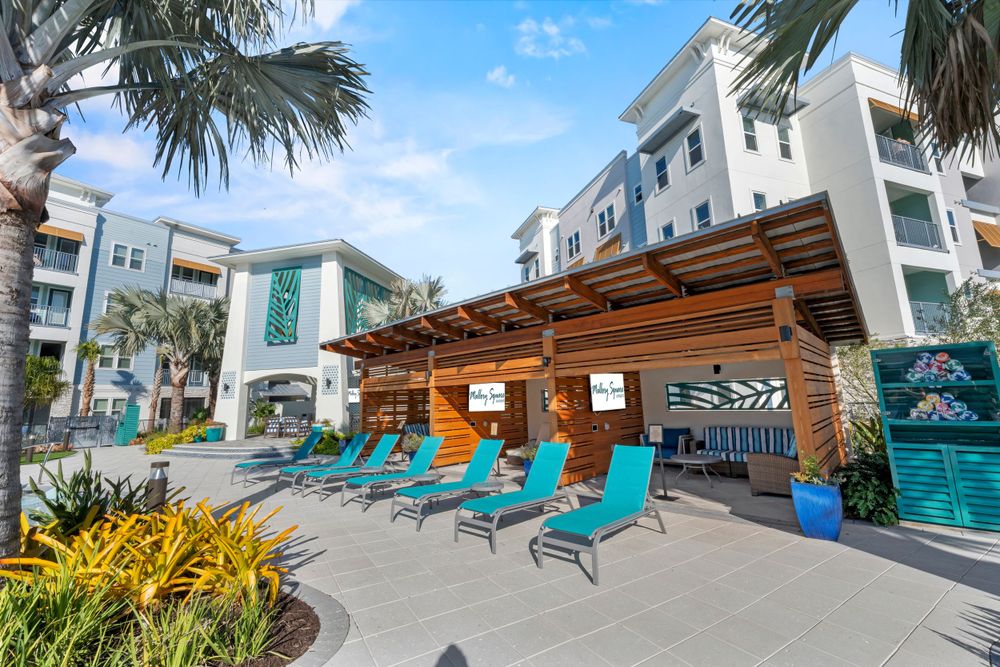 Modern apartment complex with turquoise lounge chairs and wooden pergola by the pool area surrounded by palm trees.