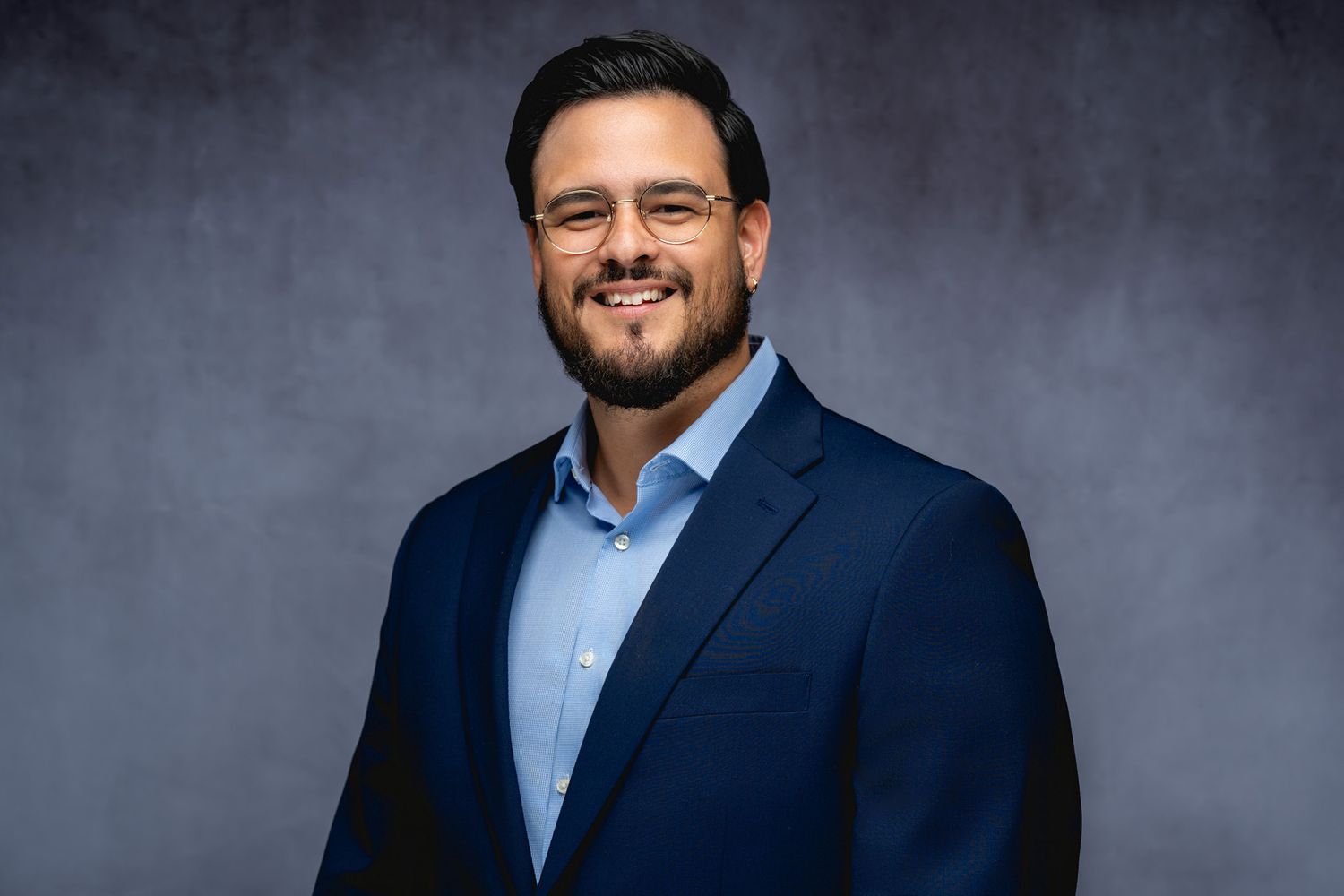 Professional headshot of a smiling person wearing a navy suit jacket and light blue button-up shirt against a gray backdrop.