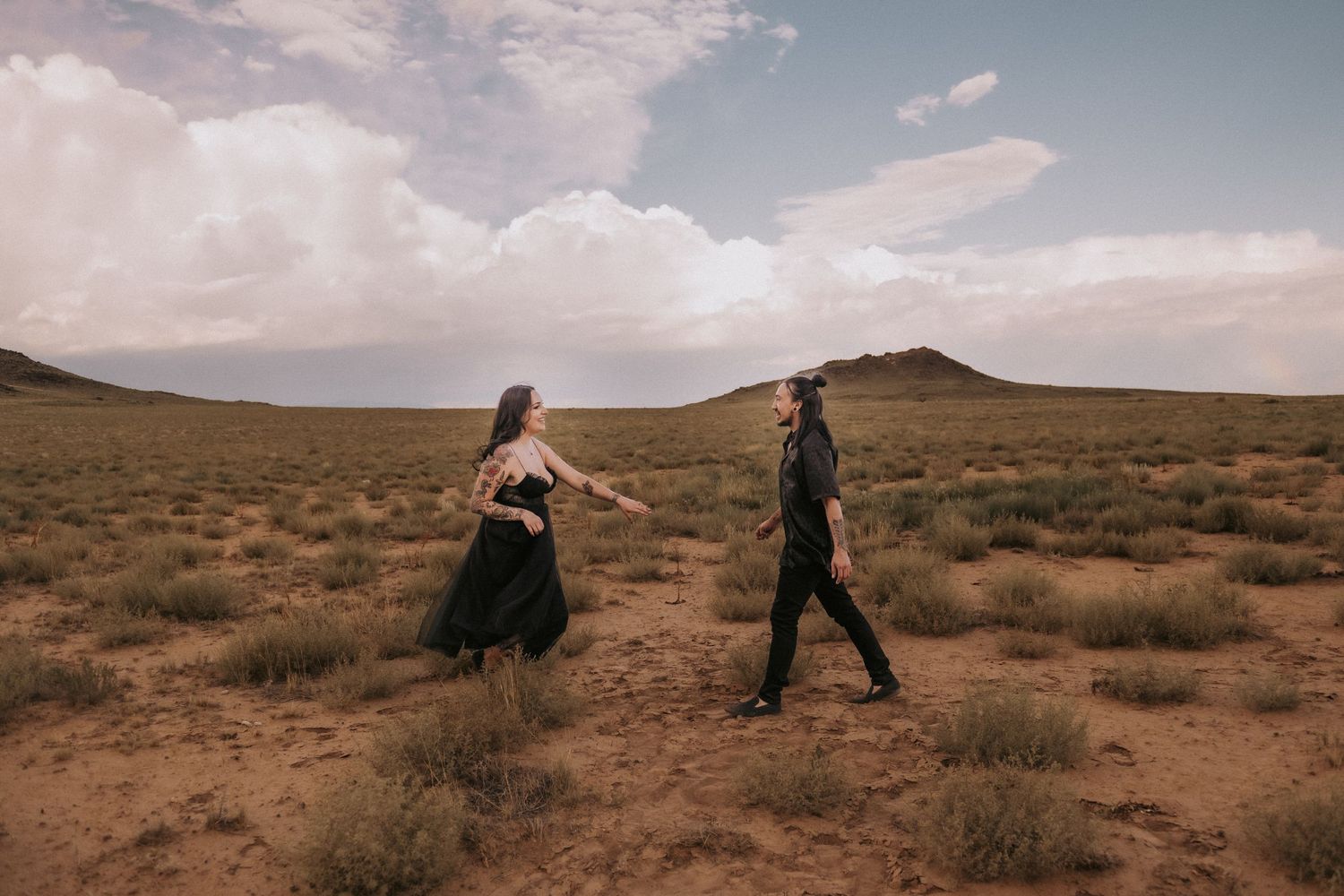 Couple walking through a desert landscape with rolling hills and cloudy skies in the background. queer wedding photographer Albuquerque.