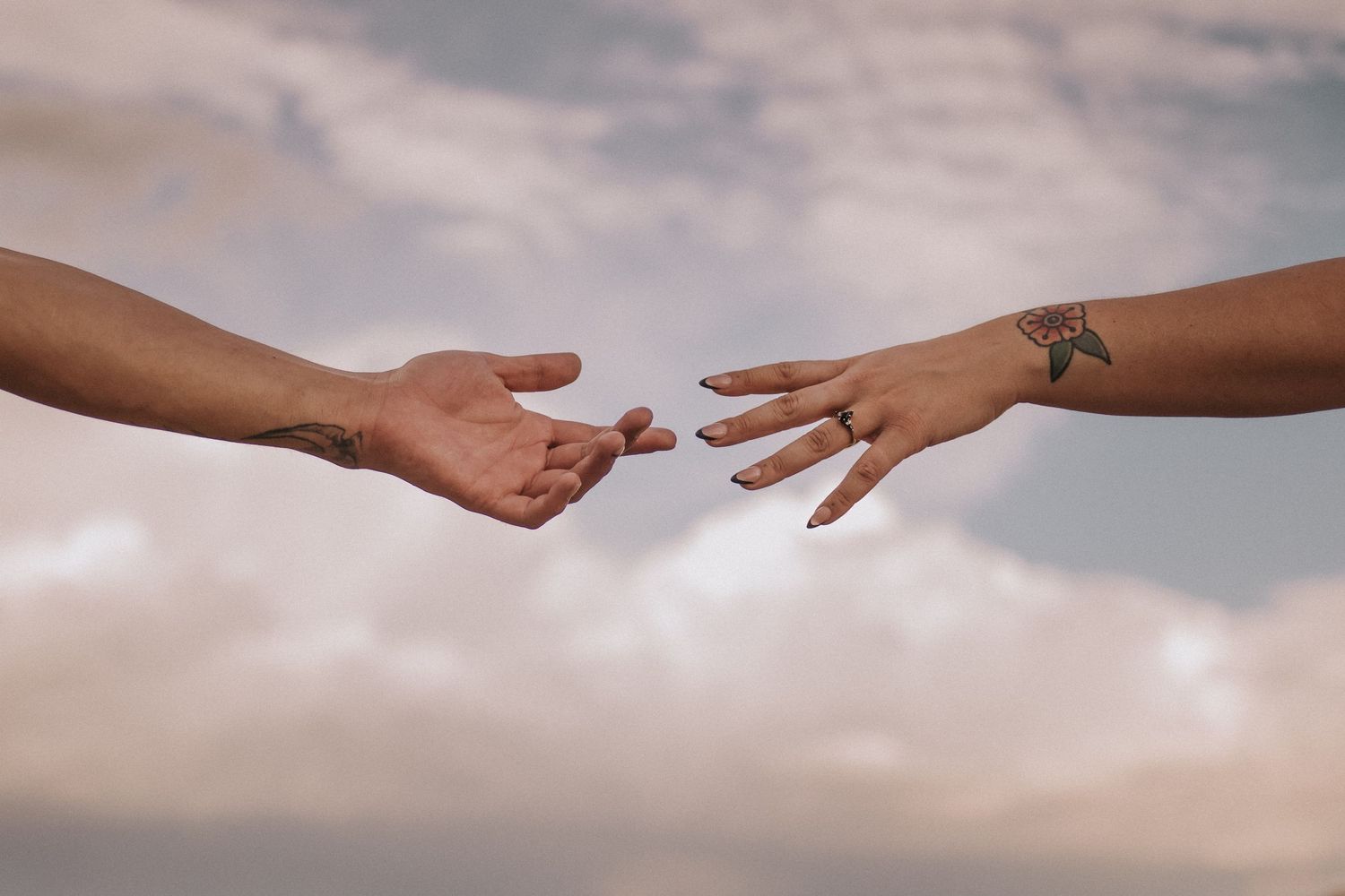 Two hands reaching towards each other against a cloudy sky backdrop with a tattoo visible on one arm. Albuquerque engagement photographer.