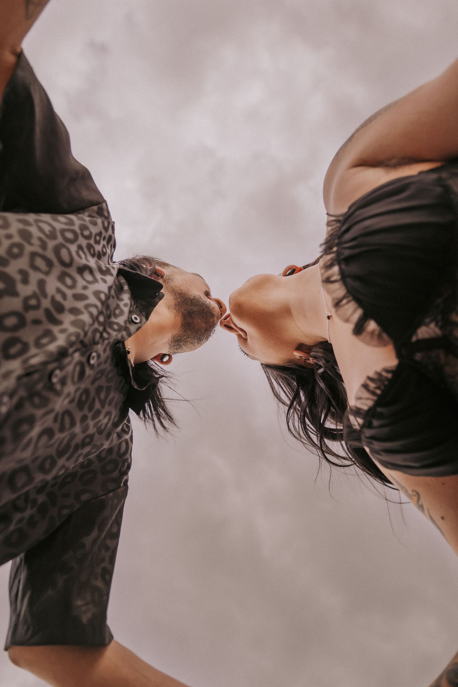 Two people with dark hair in black and leopard print clothing lean in close together against a cloudy sky in the desert in Albuquerque, NM.