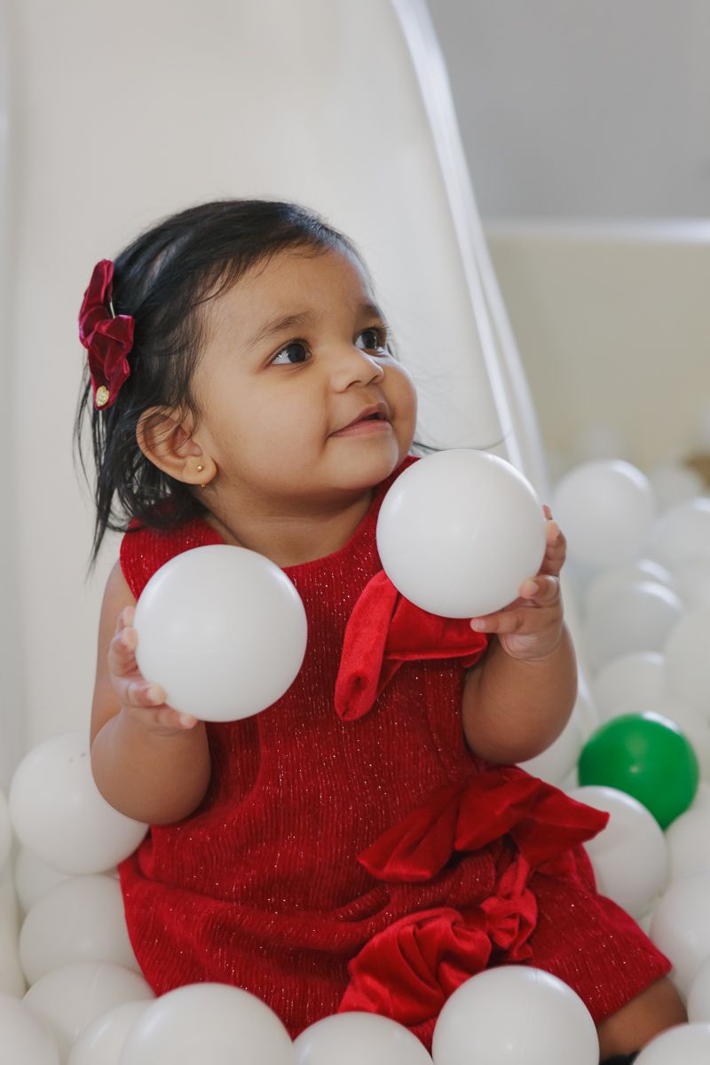 A child in a red dress with a bow playing in a white ball pit. Captured by NYC family photographer Paula Brearley