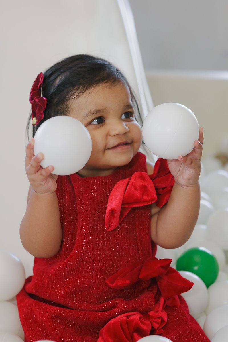 Child in a red dress with bows holding two white balls in a ball pit. Captured by NYC family photographer Paula Brearley