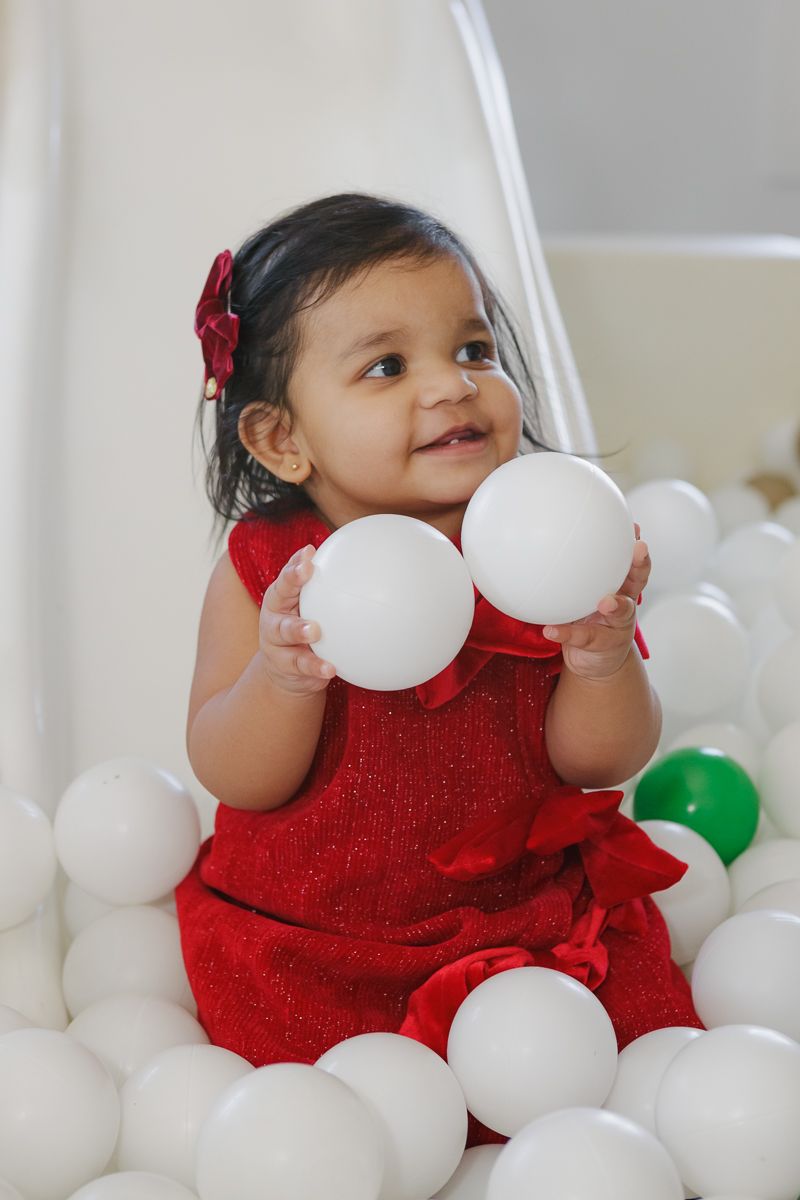 Child in a red dress with a bow playing in a white ball pit at My Gym Tribeca. Captured by NYC family photographer Paula Brearley.
