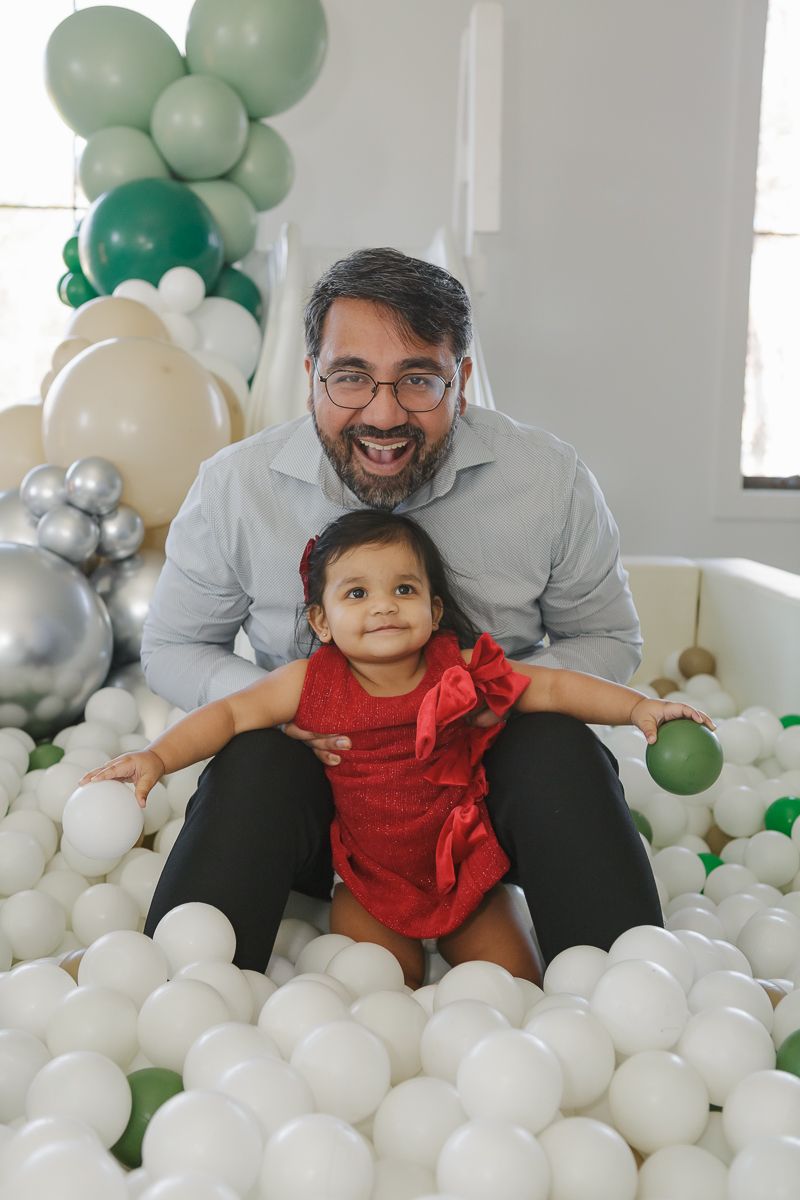 Father sitting with his daughter in a red dress with a bow in a ball pit at a festive event. Captured by NYC family photographer Paula