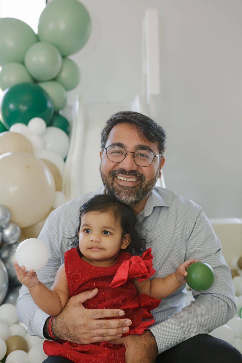 Father sitting with his daughter in a red dress with a bow in a ball pit at a festive event. Captured by NYC family photographer Paula Brear