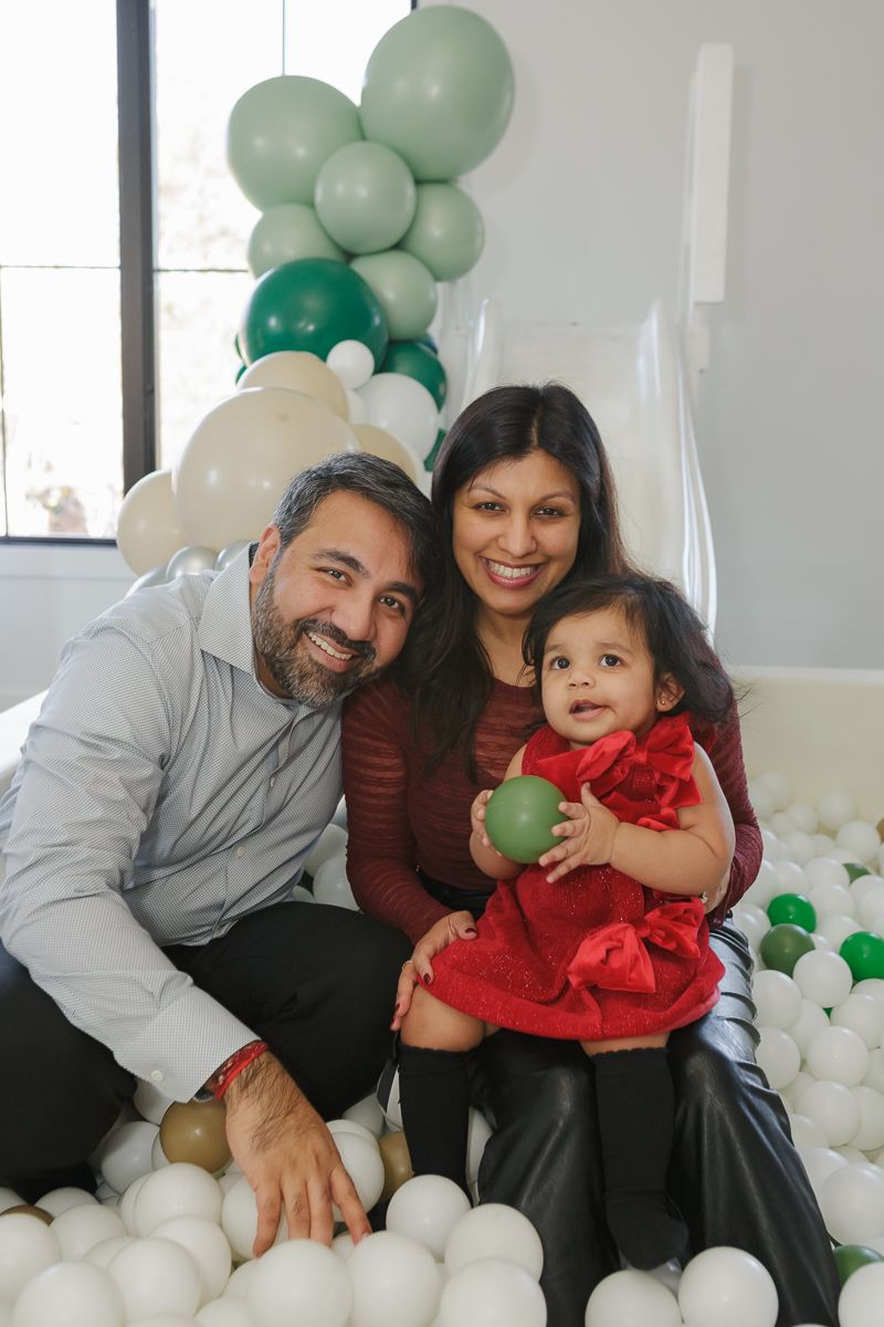 Father, mother, and child in a red dress holding a ball in a ball pit at her birthday. Captured by NYC family photographer Paula Brearley