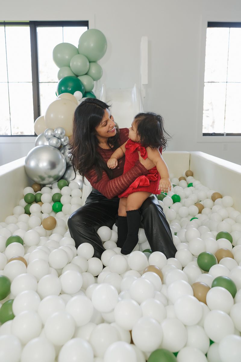 Mother and daughter in a red dress with a bow in a ball pit at a festive event. Captured by NYC family photographer Paula Brearley