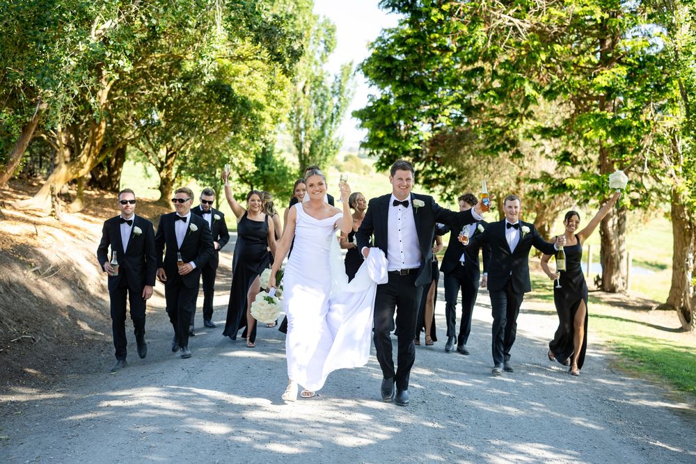 Wedding party in black tie attire celebrates joyfully on a sunlit path surrounded by lush green trees.