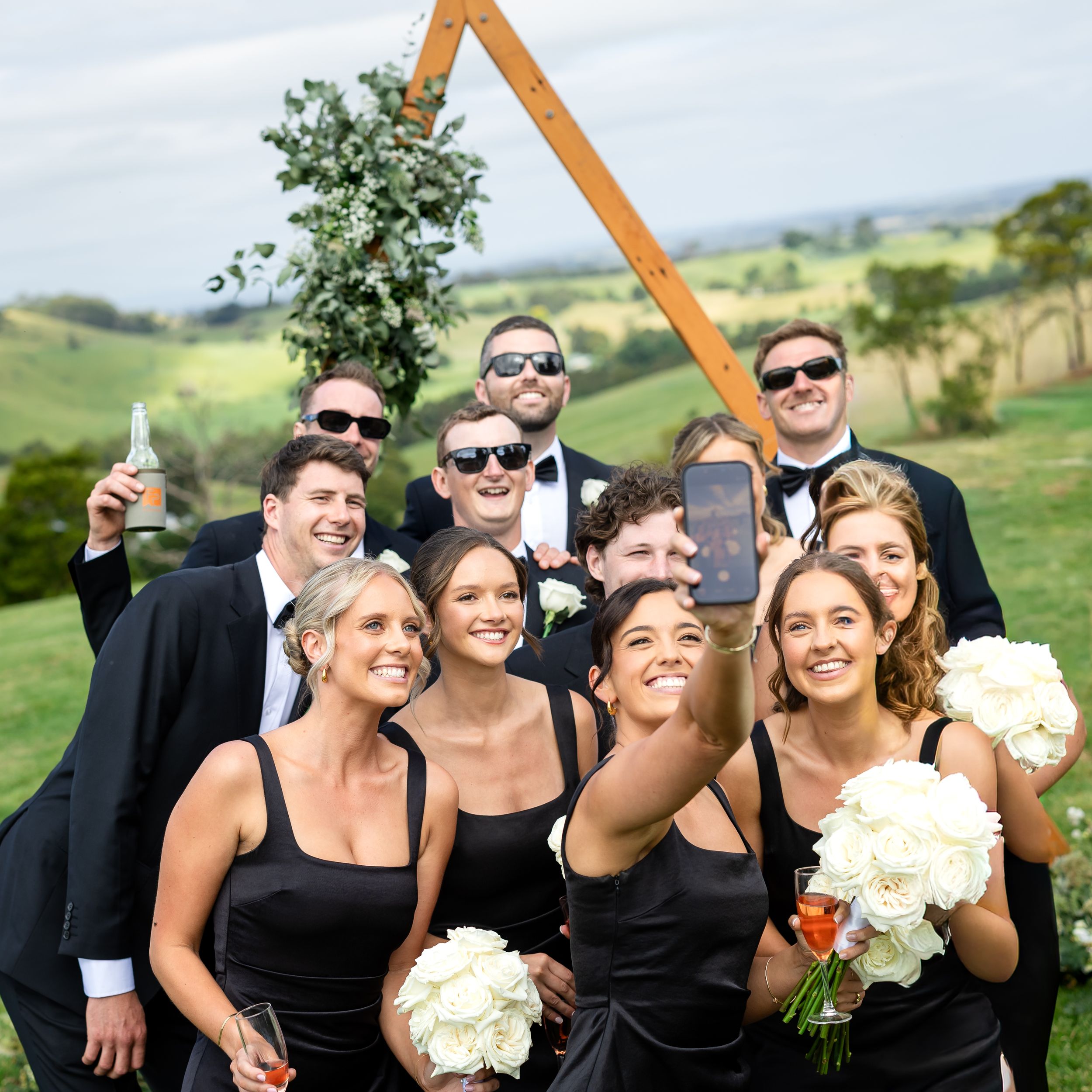 Wedding party takes a selfie outdoors, bridesmaids in black dresses with white bouquets and groomsmen in tuxedos.