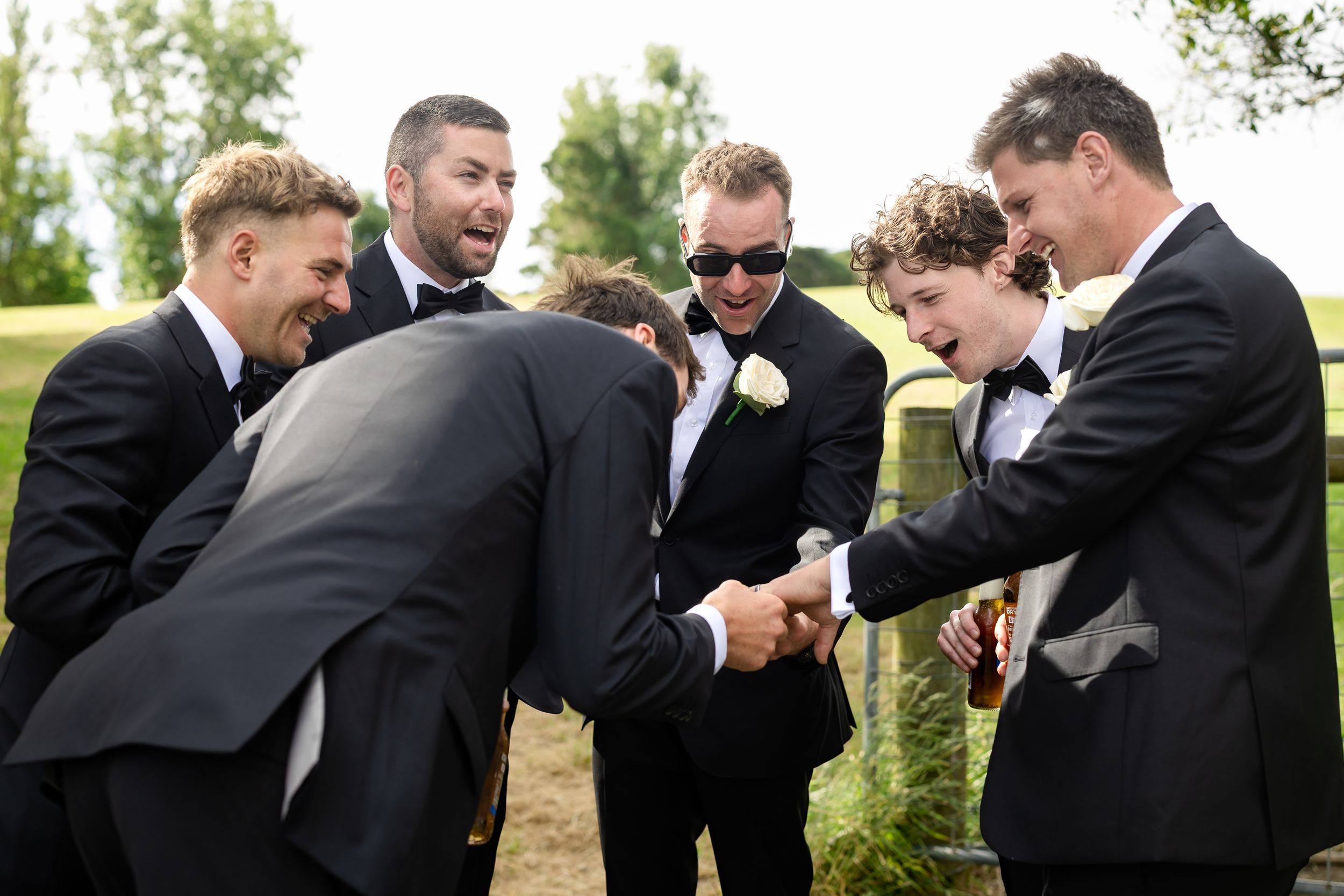 Groomsmen in black suits laughing and sharing a joyful moment together outdoors at a wedding celebration.