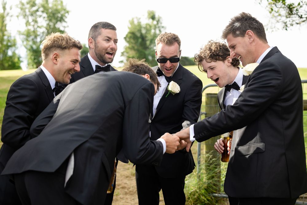 Groomsmen in black suits laughing and sharing a joyful moment together outdoors at a wedding celebration.