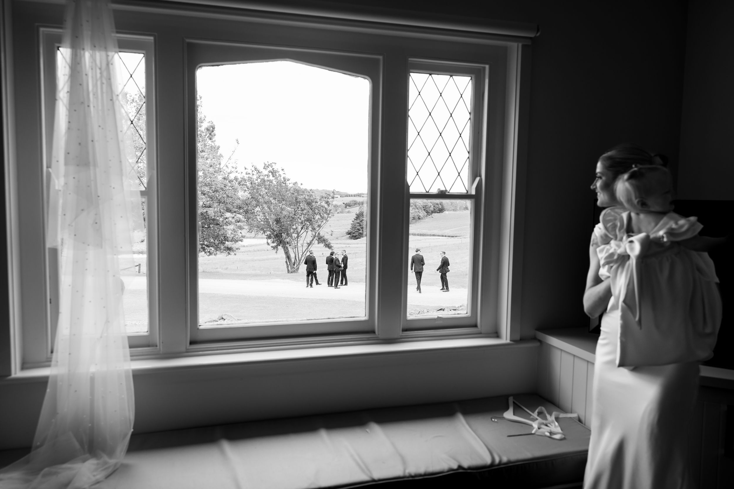 Black and white photo of a bride standing by a window, with wedding guests visible outside on a sunny day.
