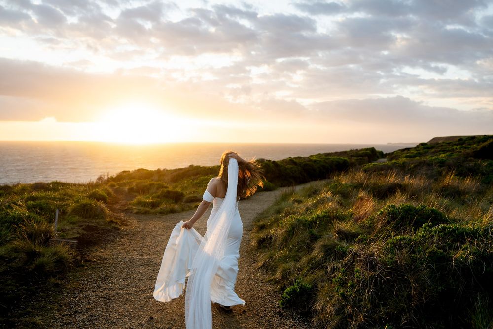 Bride in white wedding dress and veil stands on coastal path at golden sunset with ocean views.