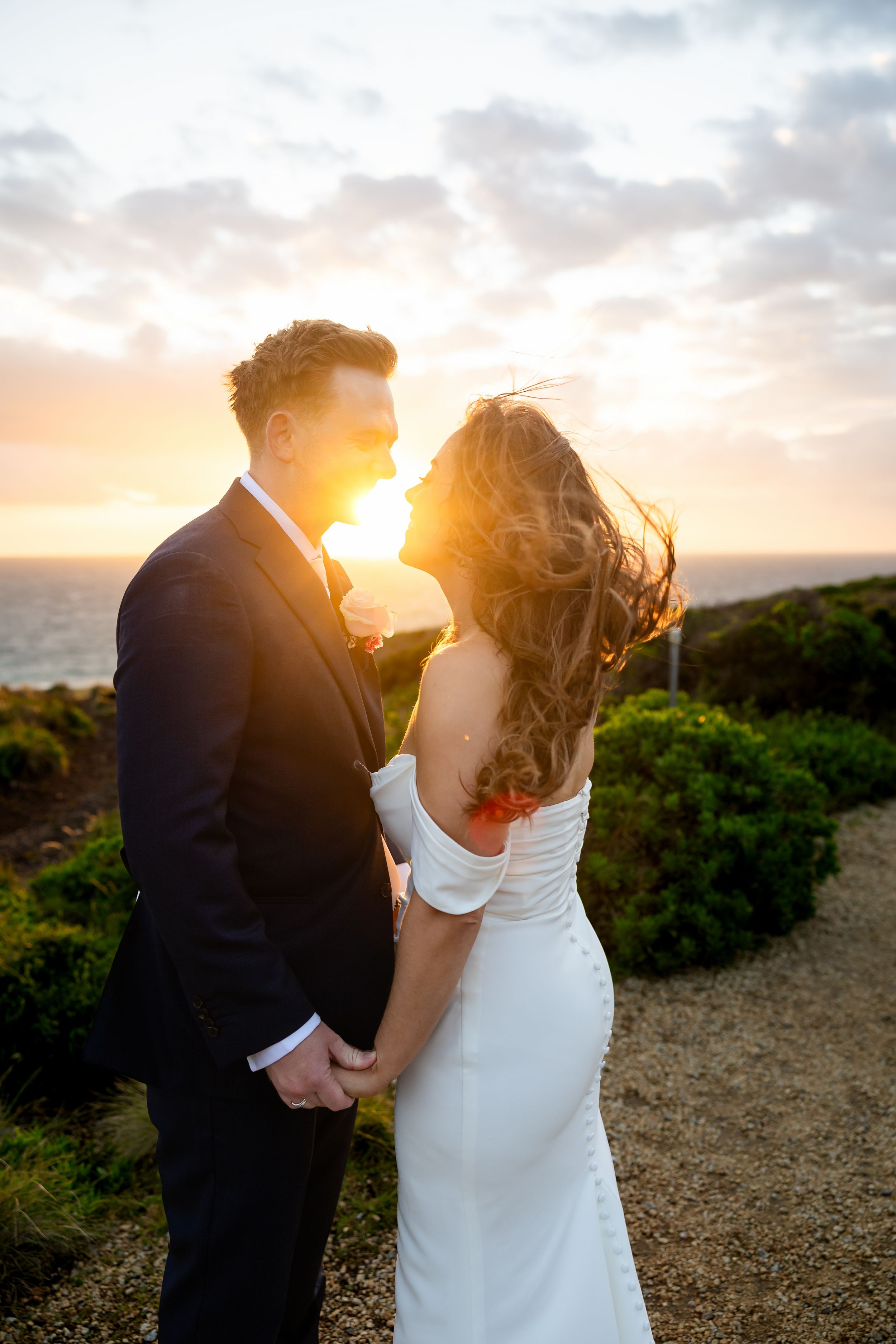 Bride and groom holding hands at sunset on coastal path, golden light glowing behind them.