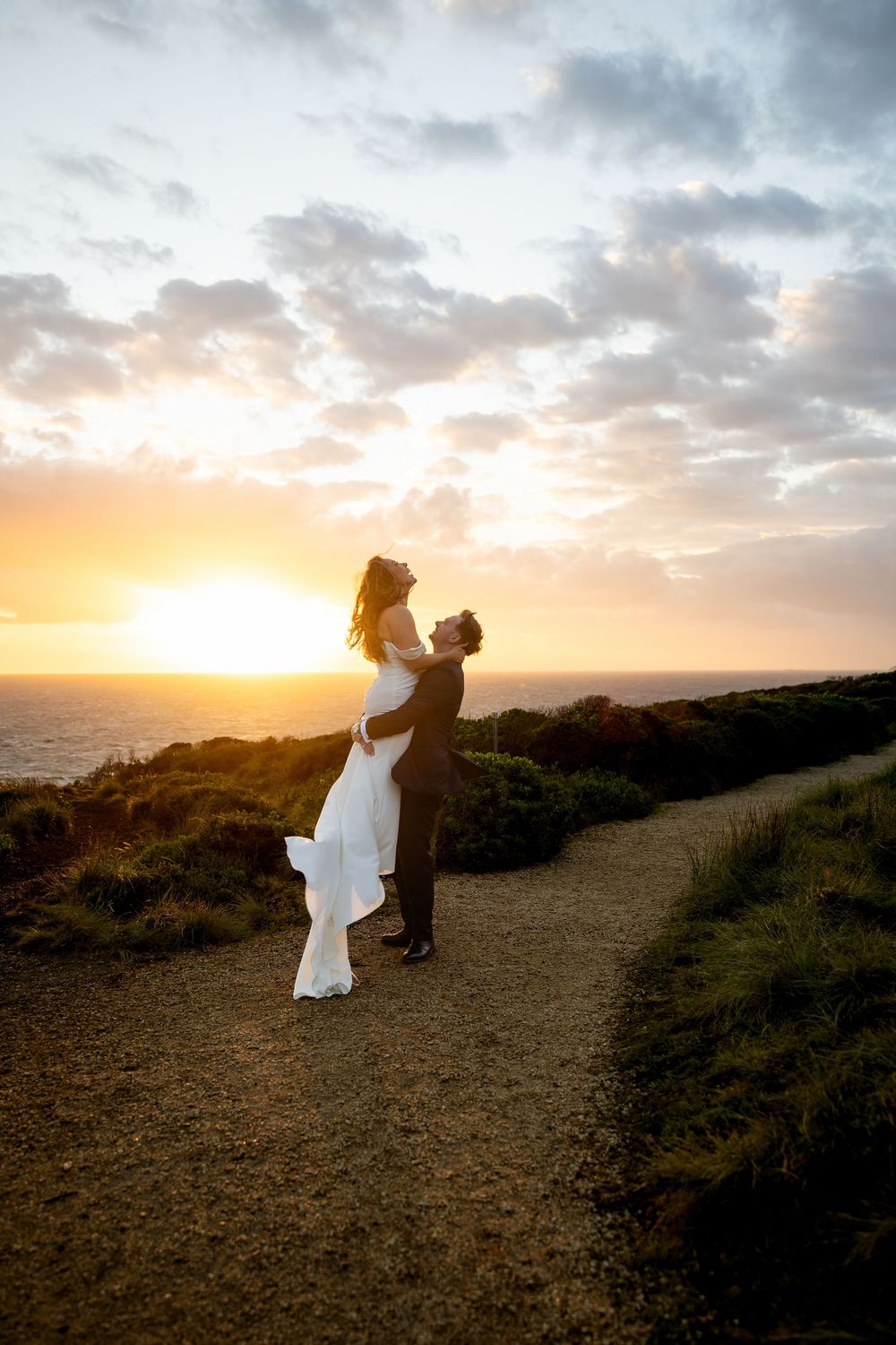 Groom lifting bride on scenic coastal path at sunset, ocean horizon glowing behind them.