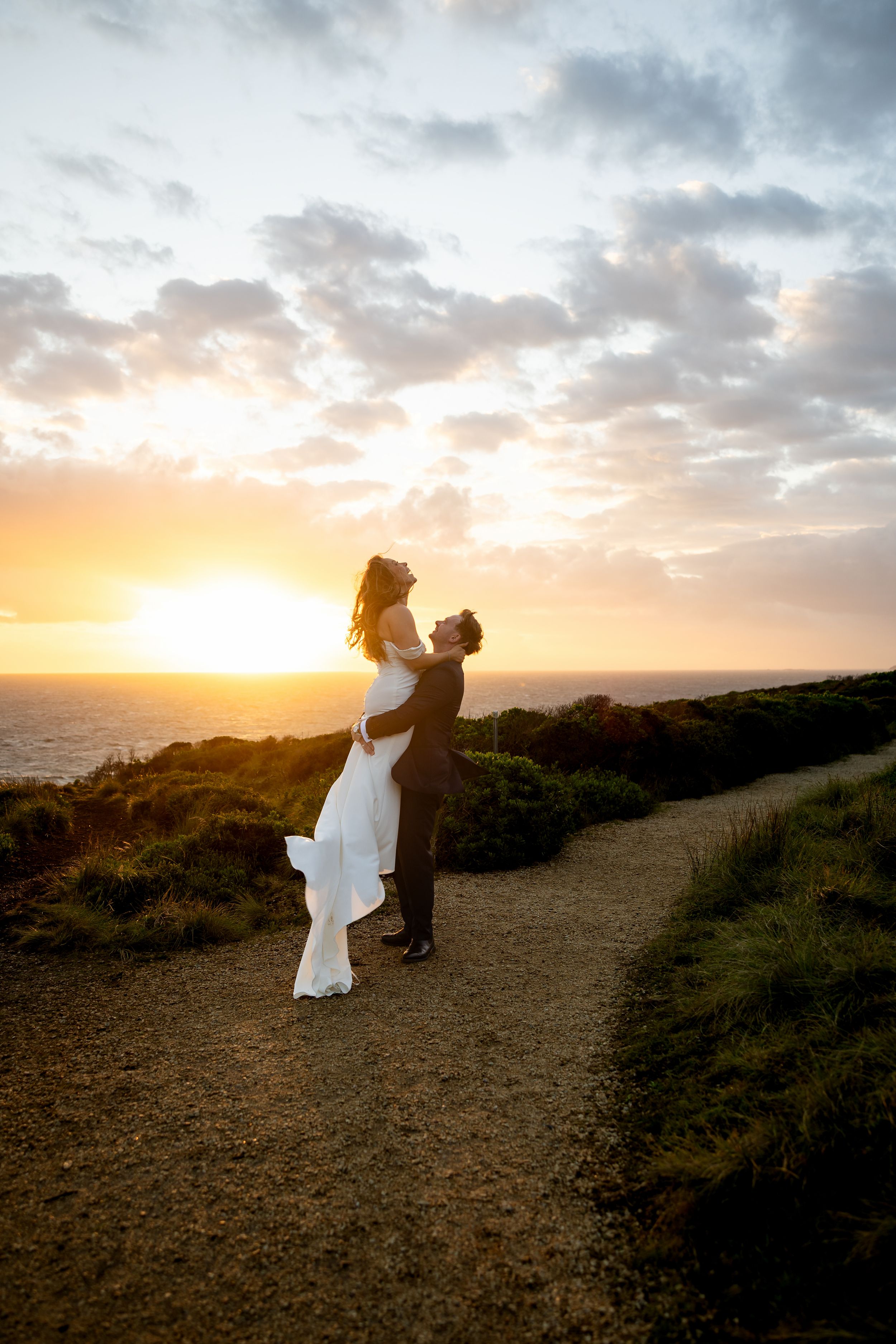 Groom lifting bride on scenic coastal path at sunset, ocean horizon glowing behind them.