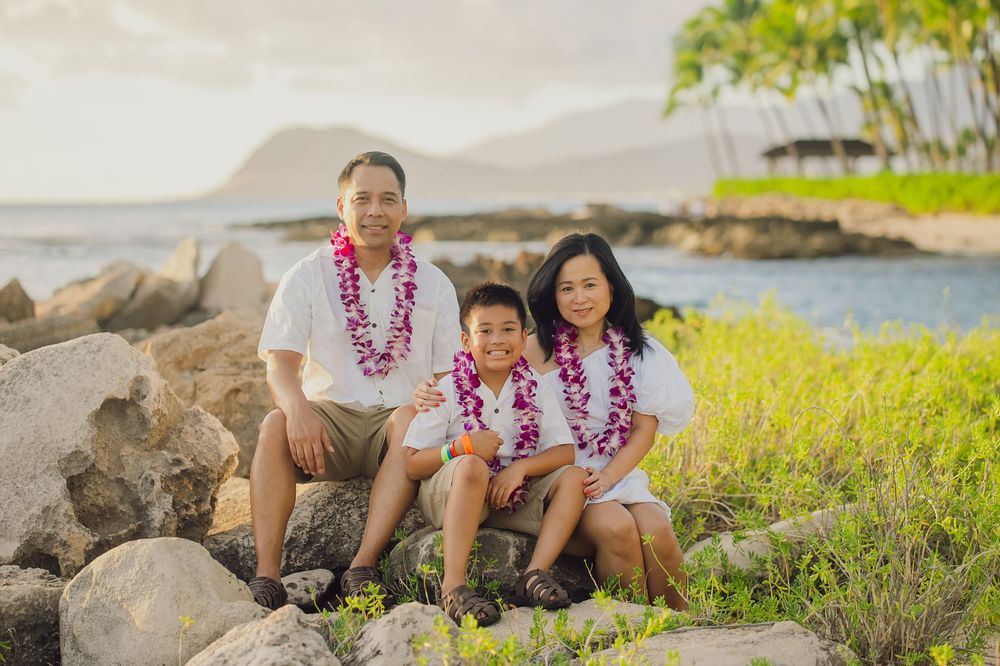 A family wearing matching white outfits and purple leis sits together on rocky beach with palm trees and ocean in Hawaii.