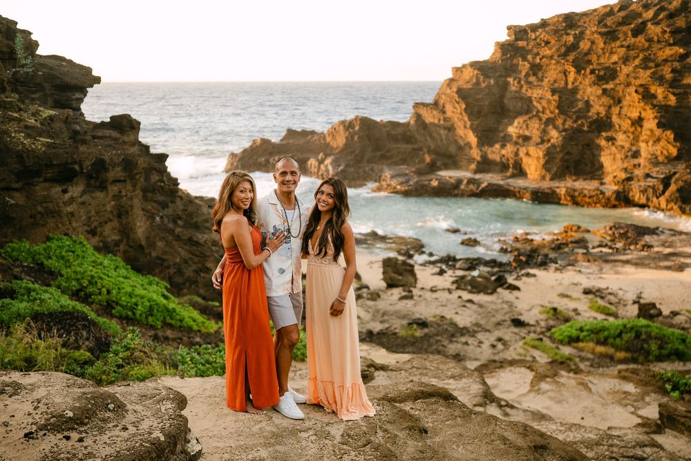 Three people posing together on rocky Hawaiian coastline at golden hour, wearing elegant maxi dresses and white suit.