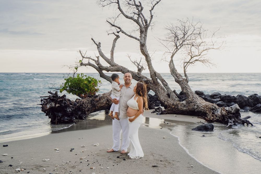 Family of three in white outfits posing on a tropical beach near driftwood trees with ocean waves in the background.
