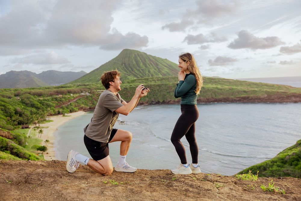 Man proposes to woman on scenic Hawaiian clifftop overlooking a lush green bay and mountain landscape.