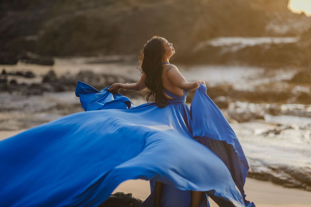 Woman in flowing blue gown on beach at golden sunset, fabric billowing dramatically in the wind.