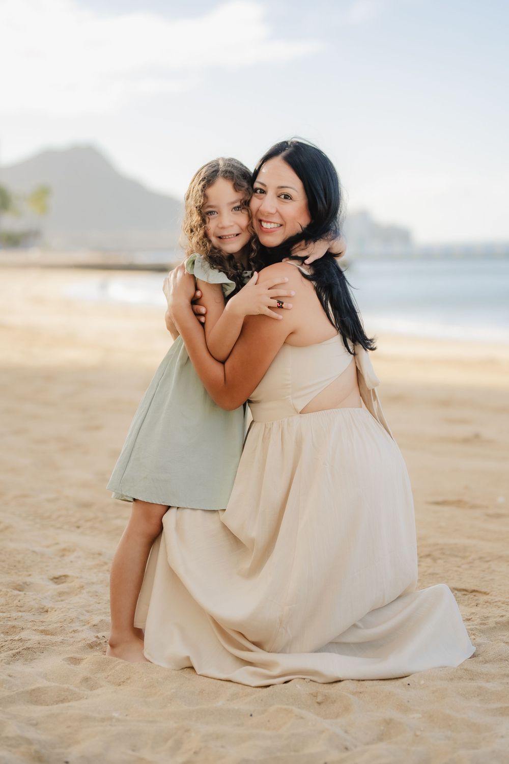 A mother and young daughter share a warm hug on a sandy beach, both smiling in light dresses with ocean views behind them.