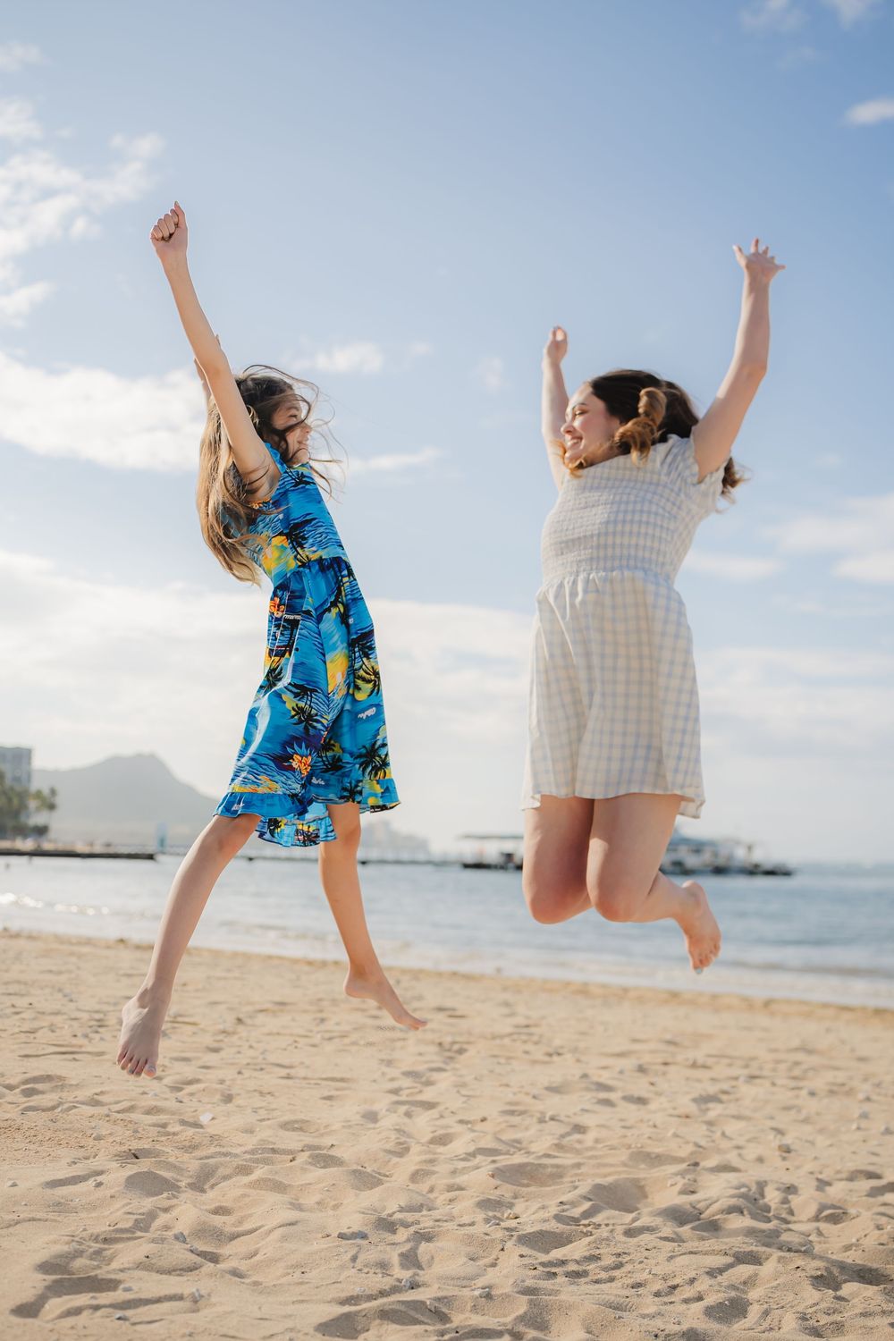 Two girls jumping joyfully on a sunny beach, wearing colorful dresses with arms raised, ocean and blue sky behind them.