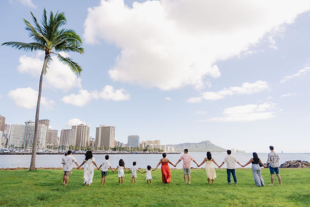 A group of people hold hands in a line stretching across a scenic waterfront with palm trees and city skyline in view.