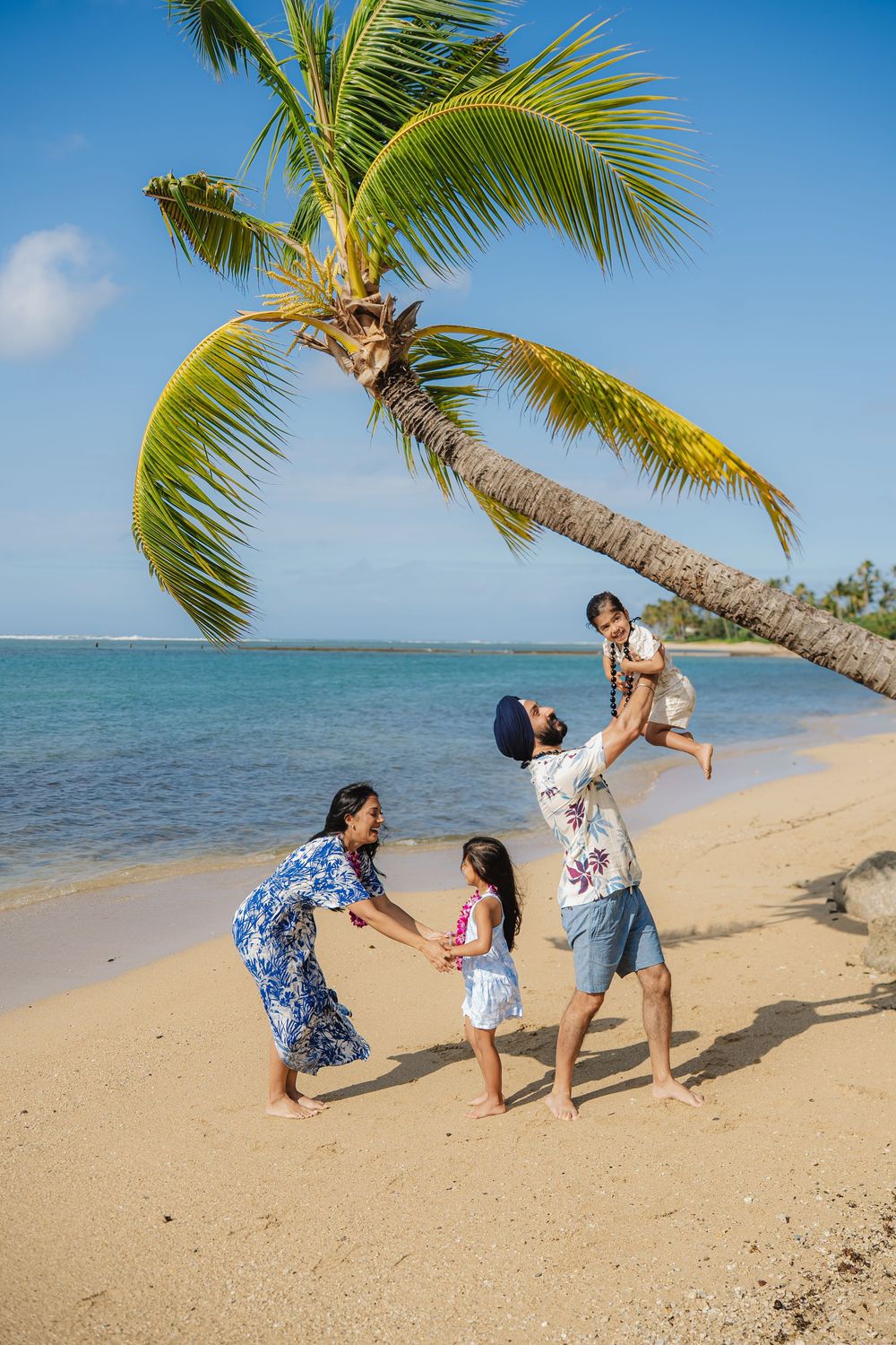 A joyful family plays on a tropical beach under a leaning palm tree with clear blue ocean waters in the background.