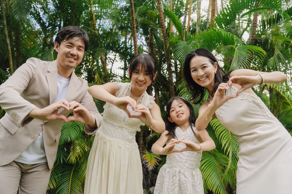 A family in coordinated beige and white outfits poses playfully together in a tropical garden setting.