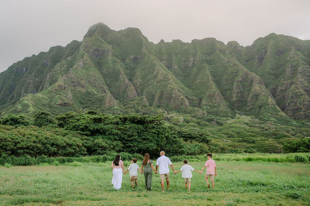 Family of six walking hand-in-hand through lush green meadow with dramatic Ko'olau Mountains backdrop in Hawaii.